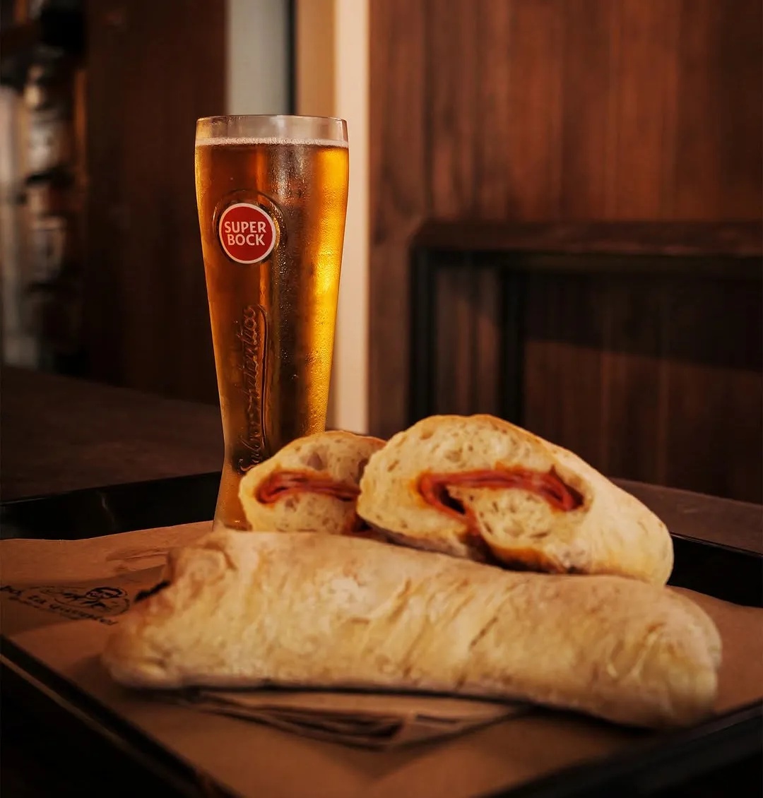 Dois pães com chouriço abertos sobre papel, com uma Super Bock